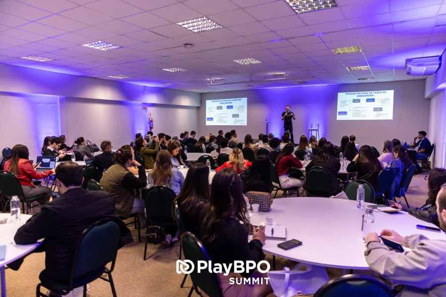 Sala de conteúdo lotada no PlayBPO Summit 2025: palestrante ao centro do palco entre dois telões, público acompanhando sentado em mesas redondas